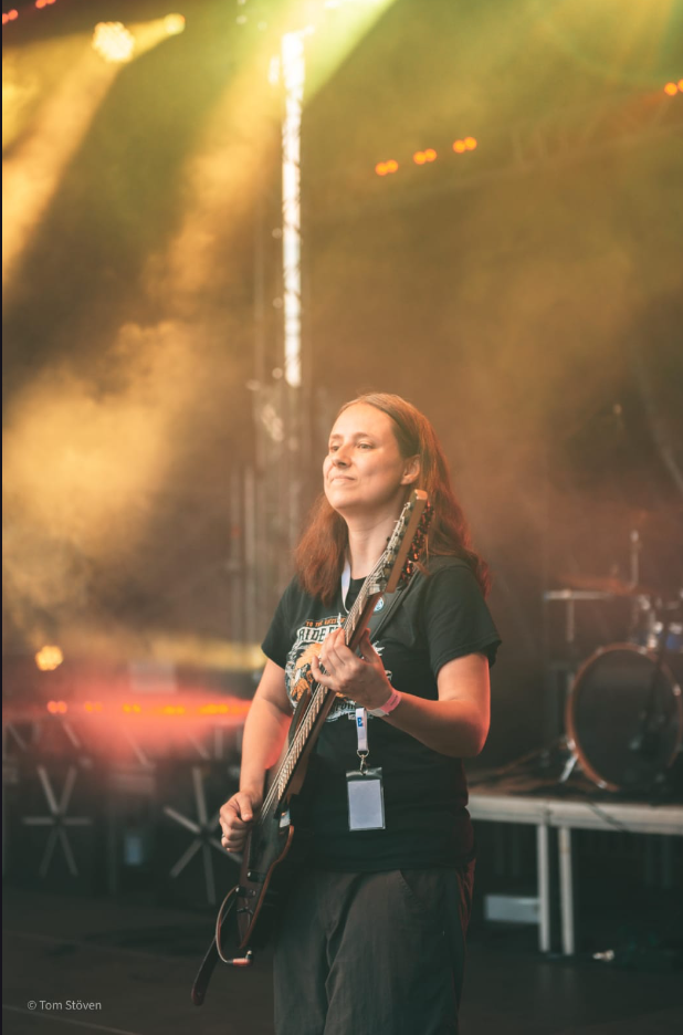 Photo of Sarina playing the bass guitar on an open air stage